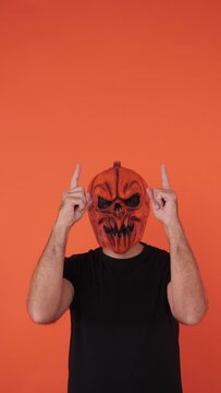 Person with pumpkin mask celebrating Halloween, pointing up with fingers, on orange background. Celebration concept, All Souls' Day and All Saints' Day.