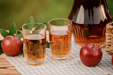 Two glasses with apple juice and basket with apples on wooden table with natural orchard background. Vegetarian fruit composition