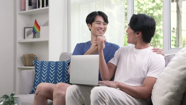 Young Asian gay couple using computer laptop in the living room and celebrating their success.