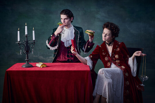 Portrait of man and woman in image of vampires over dark green background. Man eating burger, woman drinking and taking selfie