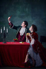 Portrait of man and woman in image of vampires taking selfie over dark green background. Mix of eras