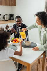 Fototapeta na wymiar african american woman pouring orange juice in glass near happy husband in suit and preteen daughter during breakfast.