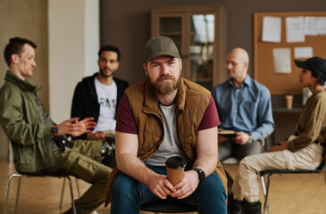 Bearded man with cup of coffee looking at camera while sitting against group of people describing their psychological problems