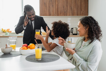 Fototapeta na wymiar happy african american girl looking at father in suit talking on smartphone during breakfast.