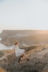 A girl in a white dress and a straw hat look at the sunset.