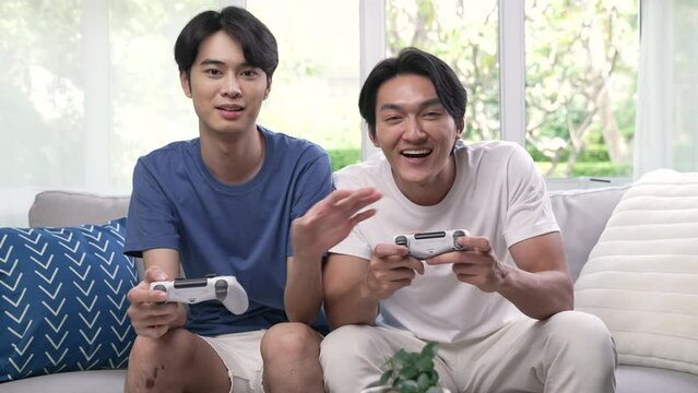 Young Asian gay couple playing video games on a couch and having fun at home. LGBT couple looking happy while spending time together at home.