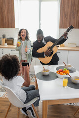 curly african american girl taking photo of happy father in suit playing acoustic guitar near mother singing in kitchen.