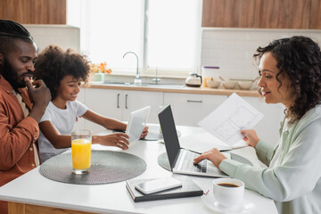 happy african american woman holding paper near laptop and family using digital tablet in kitchen.