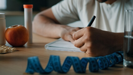 Close up unrecognizable man nutritionist at table writing prescription sport fitness diet on paper count calories healthy fresh apple and vitamins pills medicines drug and tape measure on foreground