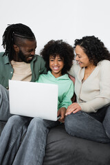 cheerful african american parents looking at preteen daughter with laptop sitting on sofa.