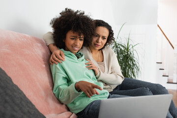 confused african american preteen girl pointing at laptop near curly mother.