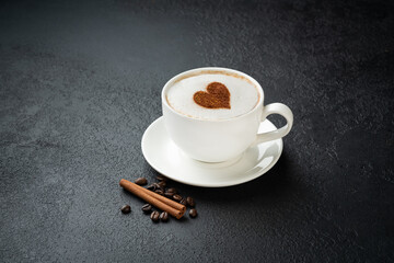 Cup of fresh capuccino with rich decorative foam with heart sign, coffee beans, cinnamon on a dark, black wooden table background