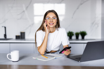 Obraz na płótnie Canvas Busy multitask woman holds a smartphone with a shoulder and typing on the laptop keyboard. Freelance woman using laptop for remote work from home, sits at the kitchen desk and has phone conversation
