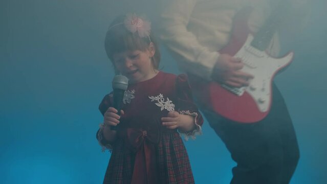 Little girl on stage in vintage dress, she sings into microphone and dances, her father plays an electric guitar. Color music is shining and smoke is billowing. Family time, creativity and hobbies