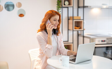 Young smiling ginger call center agent talking on phone with headset on neck and using laptop....