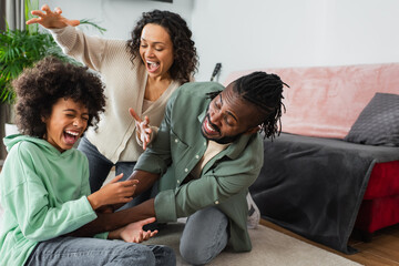 joyful african american father tickling preteen daughter near happy wife in living room.