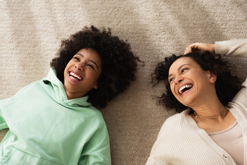 top view of happy african american girl smiling while lying on carpet with cheerful mother.