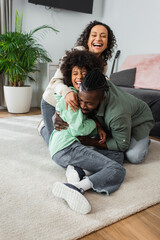 cheerful african american father tickling preteen daughter near positive wife in living room.