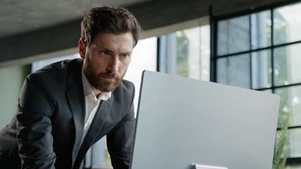 Caucasian busy bearded man adult 40s businessman employer standing in office has problem with paperwork typing data computer online check report contract work with papers and online corporate service
