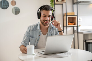 Young friendly man in good mood working as agent at call center from home while drinking coffee. Wearing headset and using laptop.