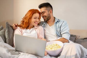 Obraz na płótnie Canvas Beautiful young happy couple watching movie together and eating popcorn while sitting on sofa.