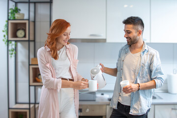 Obraz premium Cute happy young couple making tea together in the kitchen with electric kettle at home.