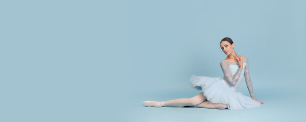 Portrait of tender young ballerina dancing, performing, sitting on floor isolated over blue studio...