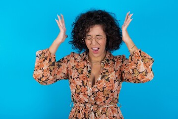 young brunette woman with curly hair wearing flowered dress standing over blue background goes...