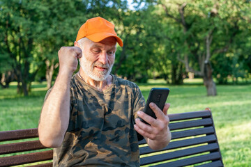Senior man in orange baseball cap with clenched fist raised up celebrates success using mobile...