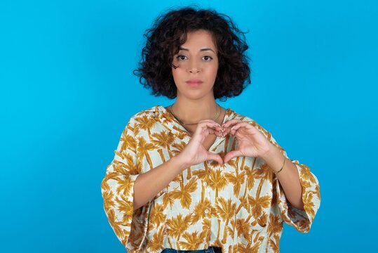 Serious young brunette woman with curly hair wearing Hawaiian printed shirt standing over blue background, keeps hands crossed stands in thoughtful pose concentrated somewhere