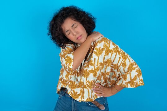 young brunette woman with curly hair wearing Hawaiian printed shirt standing over blue background got back pain