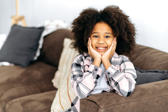 Portrait of cheerful cute african american little girl of preschool age with curly hair, in casual clothes, sits on the sofa in the living room, poses for the camera, smiling happily