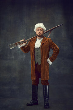 Creative portrait of retro style hunter in vintage hunting clothing with old gun isolated over dark background. Art, fashion, emotions, traditions, hobby concept