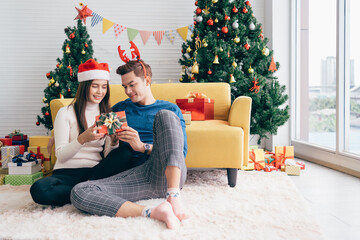 Obraz na płótnie Canvas Young beautiful happy Asian woman wearing Santa Claus hat surprises her boyfriend with a Christmas gift at home with Christmas tree in the background. Image with copy space.
