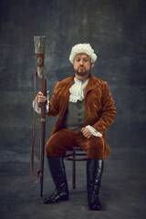 Creative portrait of retro style hunter in vintage hunting clothing with old gun isolated over dark...
