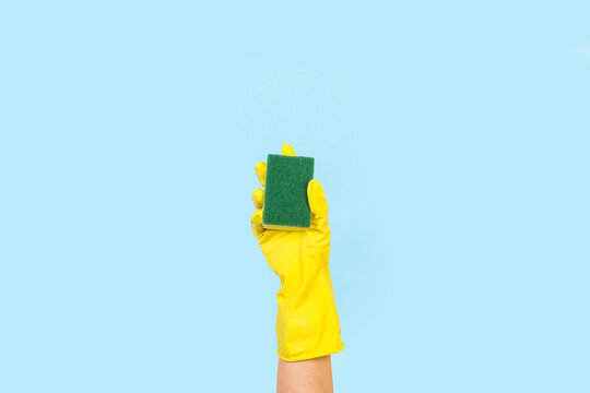 Mano con guante de goma amarillo sosteniendo una esponja de limpieza sobre un fondo celeste pastel liso y aislado. Vista de frente y de cerca. Copy space