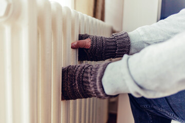 Old men's hands in knitted gloves on heating radiator at home during the day. Person heating their hands at home over a domestic radiator in winter.