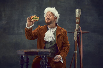Tasting burger. Vintage portrait of young man in brown vintage suit and white wig like medieval...