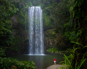 A girl in a red sweatshirt and red and white skirt stands over a powerful tropical waterfall in australia  millaa millaa falls in queensland  queensland rainforest © Lens Down Under