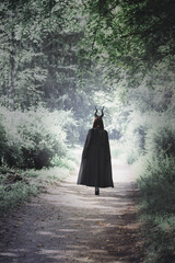 A witch with long horns in a dark cloak walks through a dark, spooky forest