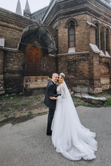 A young groom in a blue plaid suit and a cute bride in a white long dress with a diadem, a bouquet, stand embracing, near the church, the temple against the sky. wedding photography.