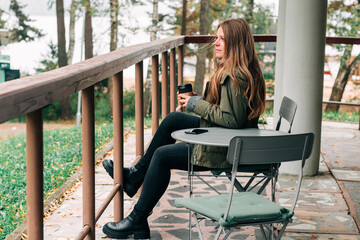 Young woman sitting on outdoor terrace in the forest drinking a cup of hot coffee. Beautiful female relax and enjoy outdoor activity lifestyle in nature on autumn travel vacation