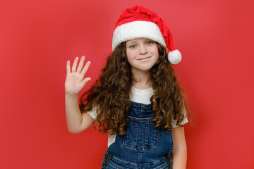 Portrait of beautiful smiling little girl in Santa hat waving hand, positive looking at camera, posing isolated over red color background wall in studio. Happy New Year 2023 holiday and Xmas concept