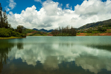Fototapeta premium Towering mountains, river with visible small island, blue and white sky, reflection visible in water, nature's gift of greenery everywhere is the way one can explain the beauty of Ooty.