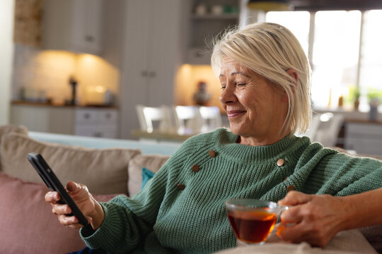 Happy Senior Caucasian Woman Sitting On Sofa In Living Room, Using Smartphone And Drinking Tea