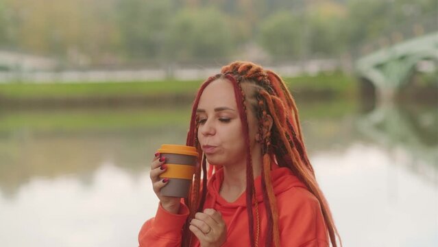 Side view of young woman drinking coffee. Female with redheads dreadlocks with a cup of coffee or tea on background of lake