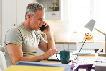 Happy caucasian man sitting at table in kitchen and using laptop and talking on smartphone