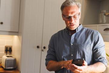 Happy caucasian man standing in kitchen and using smartphone