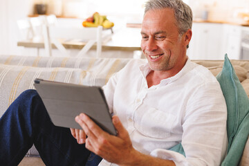 Happy caucasian man sitting on sofa in living room, using tablet