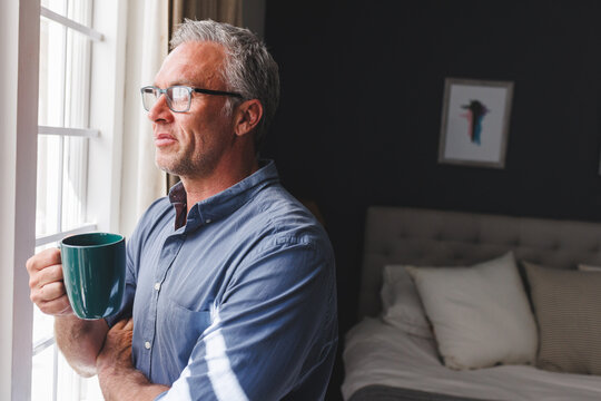 Thoughtful caucasian man looking through window and drinking coffee